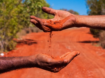 2 First Nations people in a desert. One of them is pouring red dirt into the other person's hand.