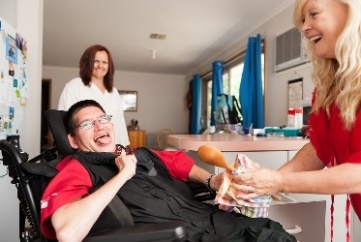 2 women help a man in a wheelchair in a household kitchen.